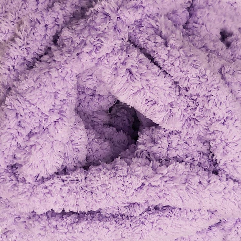 May include: Close-up of a soft, fluffy, lavender-colored blanket. The blanket has a thick, textured appearance with a dense, plush pile. The fabric is bunched and folded, creating shadows and depth.