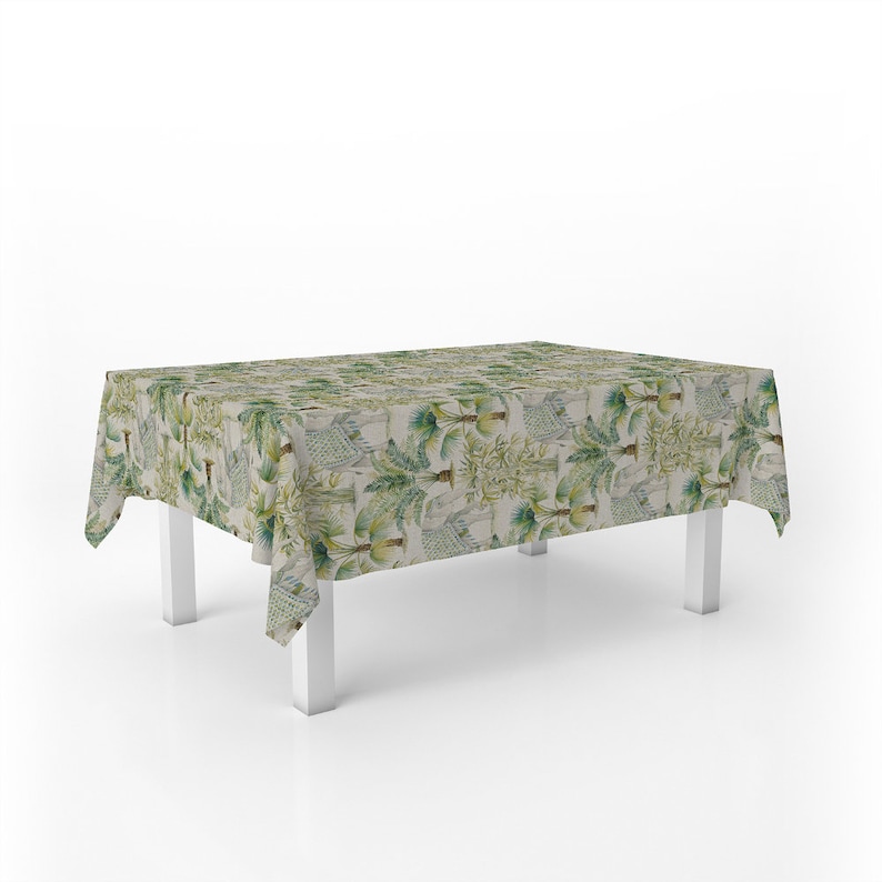 Puede incluir: Un mantel rectangular con fondo blanco y estampado tropical verde con palmeras y bamb&uacute;. El mantel est&aacute; drapeado sobre una mesa blanca con patas blancas.