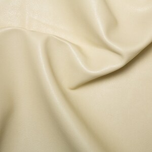 May include: Cream colored faux leather fabric with a smooth, slightly textured surface. The fabric is folded and draped, showing its soft, pliable texture.