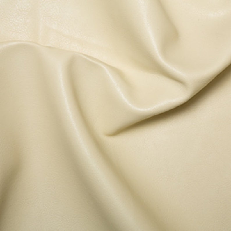 May include: Cream colored faux leather fabric with a smooth, slightly textured surface. The fabric is folded and draped, showing its soft, pliable texture.