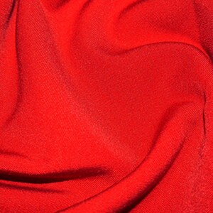 May include: A close-up of a smooth, red fabric with a slight sheen. The fabric is folded and draped, creating soft curves and folds.