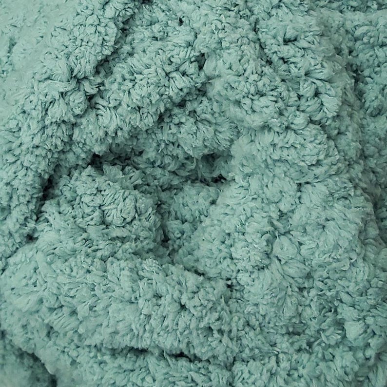 May include: Close-up of a soft, textured fabric in a muted teal color. The material has a plush, looped appearance, suggesting a cozy texture. The fabric's surface is densely packed with small, irregular loops, creating a tactile and visually appealing surface.