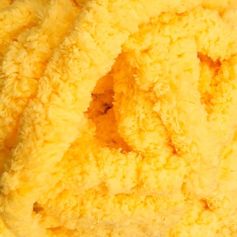 May include: Close-up of a pile of soft, yellow yarn. The yarn has a fluffy, textured appearance, with visible strands and a dense, plush feel. The color is a bright, sunny yellow, with variations in tone creating depth and dimension.