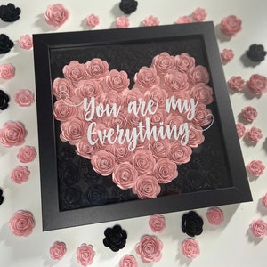May include: A framed artwork featuring a heart-shaped arrangement of pink paper roses, with the words "You are my everything" in white script. The background is black roses. Scattered around are additional pink and black paper roses.