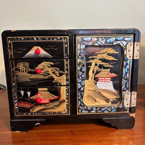 May include: Black lacquered jewelry box with three drawers and a hinged door. The box features gold and mother-of-pearl inlay depicting Japanese landscapes with pagodas, trees, and mountains. Red accents highlight key details.