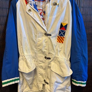 May include: A reversible jacket with a cream-colored body, blue sleeves, and a red hood. The jacket features a nautical flag patch and a patterned lining. The sleeves have green and white striped cuffs. The jacket has a zipper and toggle closures.