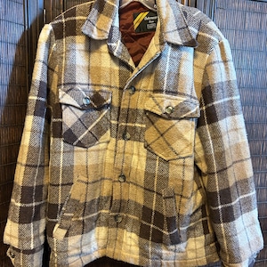 May include: A vintage plaid jacket in shades of brown, tan, and cream. The jacket features a classic button-up design with two chest pockets and a collared neckline. The interior lining is a dark brown color.