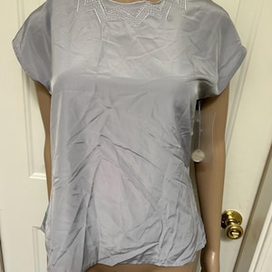 May include: A light grey, short-sleeved blouse with a round neckline. The neckline features a decorative white embroidered design. The blouse is made of a silky material and has a slightly loose fit.