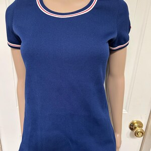 May include: A navy blue, short-sleeved shirt with a ribbed texture. The shirt features a round neckline and sleeve cuffs trimmed with white and red stripes. The shirt is displayed on a mannequin.