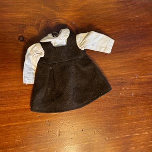 Könnte beinhalten: Ein Puppenkleid bestehend aus einem dunkelbraunen ärmellosen Trägerkleid mit ausgestelltem Rock und einem angenähten weißen Langarmhemd mit abgerundetem Kragen. Das Kleidungsstück hat ein schlichtes Oberteil und einfache Ärmel, die einen deutlichen Kontrast zwischen dem dunkelbraunen Stoff und dem weißen Hemd bilden.