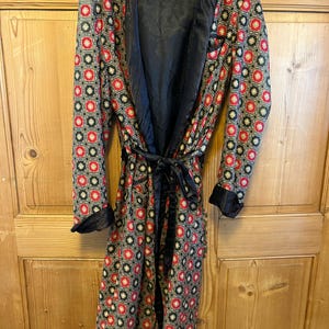 May include: A patterned robe with a black satin lining and trim. The robe features a repeating geometric design in red, black, and gold. It has a matching tie belt and long sleeves with black cuffs. The robe is hanging on a wooden door.