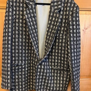 May include: A black and white patterned blazer with a notched lapel and two front pockets. The blazer has a textured weave and a striped lining. The jacket is hanging on a black hanger.