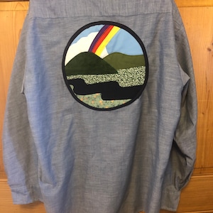 Op de afbeelding: Een lichtblauw overhemd met lange mouwen en knopen met een grote, ronde, aangebrachte patch op de rug. De patch toont een regenboog boven een groene heuvel en een rivier, alles omringd door een donkerblauwe cirkel.