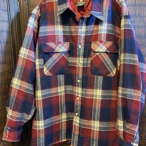 May include: A red, navy, and cream plaid flannel shirt with two chest pockets. The long-sleeved shirt has a button-down front and cuffs. The collar and inner lining are a solid burgundy color. The shirt is hanging on a hanger.