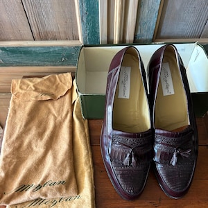 May include: A pair of burgundy leather loafers with textured detailing and tassel accents. The shoes are displayed with a shoe box and a tan dust bag. The brand name "Mezlan" is visible on the shoe's insole.