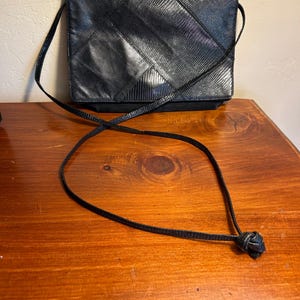 May include: Black crossbody bag with a geometric pattern. The bag features a long, thin strap and a textured surface. The bag is rectangular and appears to be made of leather or a leather-like material.