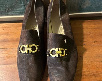May include: A pair of brown suede loafers with a gold-colored metal buckle. The shoes have a pointed toe and a low heel. The loafers are displayed on a wooden surface.