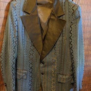 May include: A blue blazer with a black satin lapel and a patterned design. The blazer has two front pockets and three buttons on the sleeve cuffs. The jacket is hanging on a gold hanger.