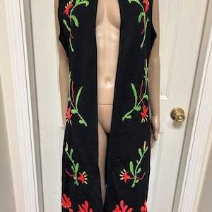May include: Black sleeveless vest with floral embroidery. The vest features a long, open front design and is adorned with red, yellow, and green embroidered flowers and leaves. The embroidery is placed along the edges and front of the vest.
