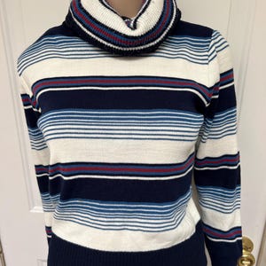 May include: A striped turtleneck sweater in shades of navy, white, and blue with a touch of red. The sweater features horizontal stripes and a ribbed collar and cuffs. The garment is displayed on a mannequin.