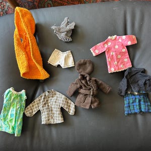 May include: A collection of miniature doll clothing including an orange hooded towel, a black and white patterned top, white shorts, a pink polka dot shirt, a brown coat, a green patterned sleeveless top, a gray patterned shirt, and a black and blue plaid skirt.