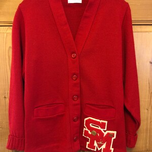 May include: Red cardigan sweater with a large white felt patch with the letters "SM" on the front. The sweater has a V-neck and button closure. The sweater has two pockets on the front.