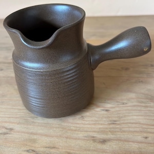 Pode incluir: Um creamer de cerâmica marrom com uma superfície texturizada e uma alça.