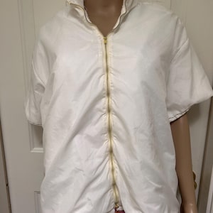 Puede incluir: Chaqueta blanca de manga corta con cierre de cremallera dorada. La chaqueta tiene un cuello alto y un diseño ligeramente abombado. La tela parece ligera y texturizada, adecuada para combinar. Estilo casual.
