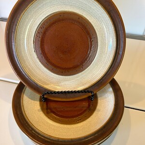 May include: Set of three ceramic plates with a concentric design. The plates feature a dark brown rim, a beige outer ring, a light beige middle ring, and a dark brown center. The plates are stacked on a black metal plate stand.