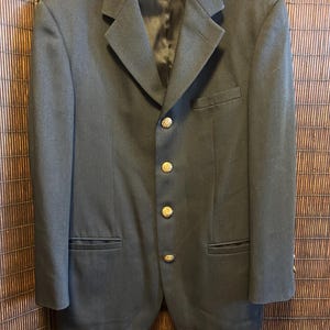 May include: A dark gray blazer with gold-colored buttons. The blazer has a classic design with a notched lapel, two front pockets, and a single-breasted closure. The blazer is hanging on a black hanger.