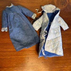 May include: A blue long-sleeved dress with white lace trim on the cuffs and collar, alongside a light blue quilted coat with a dark blue interior lining and a visible white fabric tag near the collar area, both designed for a doll.