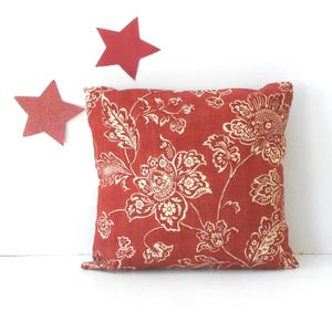 May include: Square decorative pillow with a red floral pattern and off-white accents. The pillow features a detailed botanical design. Two red star-shaped decorations are in the background.
