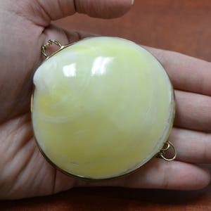 May include: A round, pale yellow object with a gold-colored metal rim and small loops. The object has a smooth, glossy surface with a marbled appearance. The item is held in a hand, suggesting its size and portability.