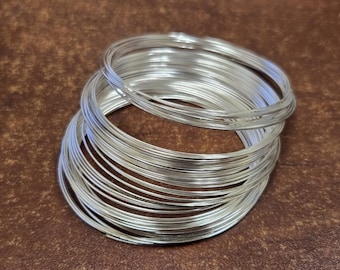 May include: A bundle of silver-colored wire coils, tightly wound and stacked. The wire has a shiny, metallic appearance, reflecting light. The coils are arranged in a circular pattern, creating a textured surface. The background is a brown, textured surface.