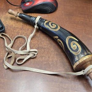 May include: A curved black horn powder flask featuring light brown spiral engravings. The object has a wooden stopper at the wide end and a smaller wooden tip at the narrow end, with a light tan leather strap attached for carrying or suspension.