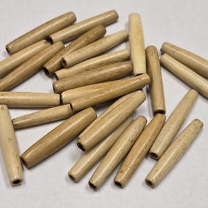Op de afbeelding: Een stapel natuurlijke houten kralen, elke kraal is lichtbruin van kleur en heeft een cilindrische vorm. De kralen zijn ongeveer 2,5 cm lang en 0,6 cm in diameter.