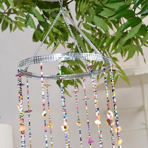 May include: A colorful beaded wind chime with shells hanging from a silver metal ring. The wind chime is suspended from a silver chain.