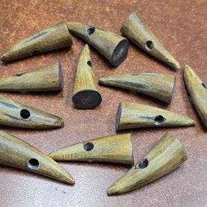May include: A collection of horn toggle buttons in various sizes and shapes. The buttons are a light brown color with dark brown and black accents. Each button has a hole for sewing. The buttons are arranged on a brown surface.