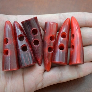 May include: Six red, horn-shaped buttons with three holes each. The buttons have a glossy finish and a dark red grain pattern. They are arranged on a light brown surface.