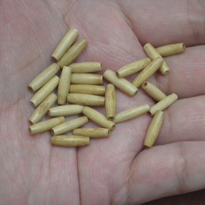 May include: A collection of small, light-colored wooden beads, cylindrical in shape with rounded ends and a hole through the center. The beads are scattered in a person's open hand, showcasing their natural wood grain.