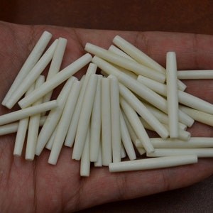 May include: A collection of approximately 40 white bone beads, each about 1 inch long and 1/4 inch in diameter. The beads are smooth and polished, with a slightly irregular shape. They are arranged in a pile on a hand.
