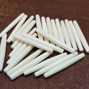 May include: A collection of off-white, cylindrical beads with rounded ends. The beads are scattered on a brown surface. The smooth, polished texture of the beads is visible in the image. These beads could be used for jewelry making or other craft projects.