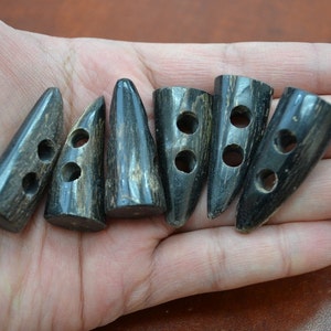 May include: Six dark brown, horn-shaped toggle buttons with two holes each. The buttons are arranged in a row, showing variations in shape and grain. Ideal for coats, jackets, or other garments.