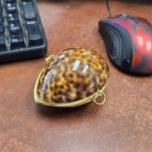 May include: A decorative, oval-shaped trinket box with a gold-colored frame and a spotted, brown and tan shell-like top. The box is on a brown surface, next to a black keyboard and a red and black computer mouse.