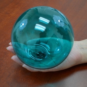 May include: A teal blue glass sphere, approximately 4 inches in diameter, is held in a person's hand. The sphere is smooth and reflective, with a slight swirl pattern visible inside.