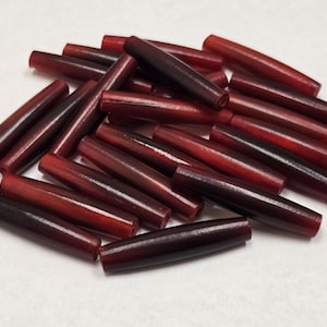 Può includere: Un mucchio di perline cilindriche lisce di colore rosso scuro e bordeaux. Le perline sono fatte di un materiale lucido, probabilmente legno o plastica.