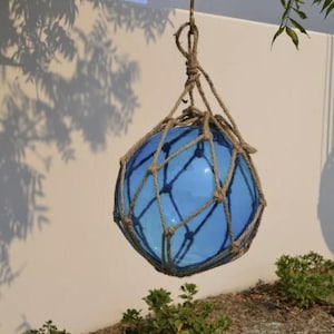 May include: A hanging blue glass float, encased in a woven rope net. The float is suspended from a knotted rope, with shadows of leaves and branches cast on the white wall behind it. Green bushes are at the base.