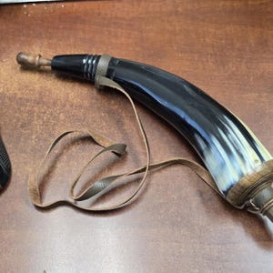 May include: A curved, polished black and white horn powder flask featuring a wooden stopper at the narrow end and a wooden base cap at the wider end, equipped with a thin, light brown leather carrying strap attached to the body.