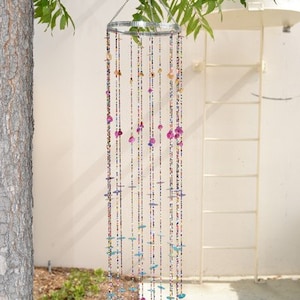 May include: A colorful beaded wind chime with pink, blue, and white beads hanging from a metal ring. The wind chime is hanging from a tree branch.