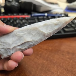 Peut inclure: Une pointe de projectile en pierre allongée de couleur gris clair, dotée d'une extrémité pointue et d'une surface texturée et écaillée. L'objet présente une forme triangulaire effilée avec des bords taillés visibles sur les côtés.